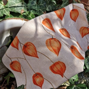 May include: A beige fabric with a repeating pattern of orange Chinese lanterns. The fabric is folded and lying on a bed of green leaves.