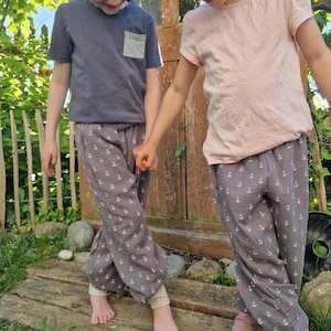 May include: Two children wearing matching gray pants with white anchor patterns and light-colored cuffs. One child wears a gray t-shirt with a pocket, the other a pink t-shirt. They stand on a wooden walkway in a garden.