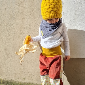 Op de afbeelding: Een kind draagt ​​een gele gebreide muts met een pompon, een wit shirt, een grijs-witte bandana en een rode corduroy broek met een bloemenpatroon. Het kind houdt een maïs kolf vast.