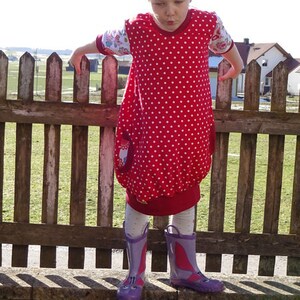 Puede incluir: Una ni&ntilde;a peque&ntilde;a con un vestido rojo con lunares blancos y un lazo rosa en su sombrero de paja. Est&aacute; de pie frente a una valla de madera y lleva botas de lluvia moradas.
