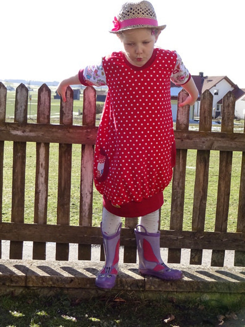 Puede incluir: Una ni&ntilde;a peque&ntilde;a con un vestido rojo con lunares blancos y un lazo rosa en su sombrero de paja. Est&aacute; de pie frente a una valla de madera y lleva botas de lluvia moradas.