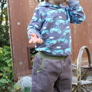 May include: A young child wearing a grey and brown striped knitted hat, a grey long-sleeved shirt with a blue and green tractor print, and grey corduroy trousers. The child is standing in a grassy area with a wooden door and a wicker basket in the background.