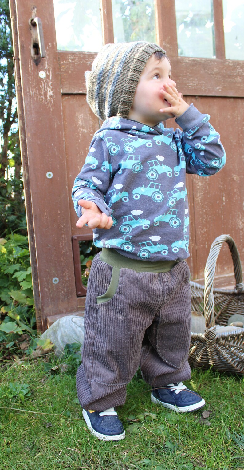 May include: A young child wearing a grey and brown striped knitted hat, a grey long-sleeved shirt with a blue and green tractor print, and grey corduroy trousers. The child is standing in a grassy area with a wooden door and a wicker basket in the background.