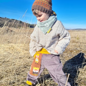 May include: A child wearing a brown and orange striped knit hat, a light green bandana, a cream colored sweater, and gray corduroy pants with a yellow waistband and a cartoon character print. The child is standing in a field of tall dry grass.