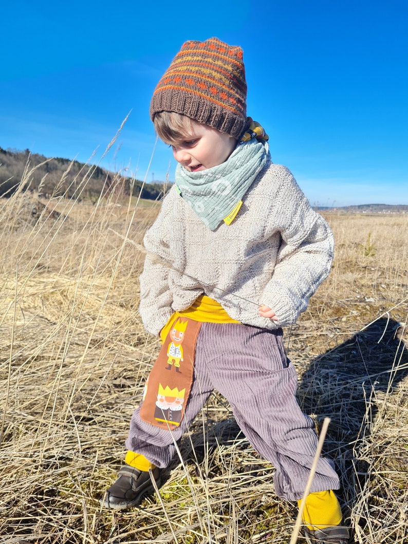 May include: A child wearing a brown and orange striped knit hat, a light green bandana, a cream colored sweater, and gray corduroy pants with a yellow waistband and a cartoon character print. The child is standing in a field of tall dry grass.