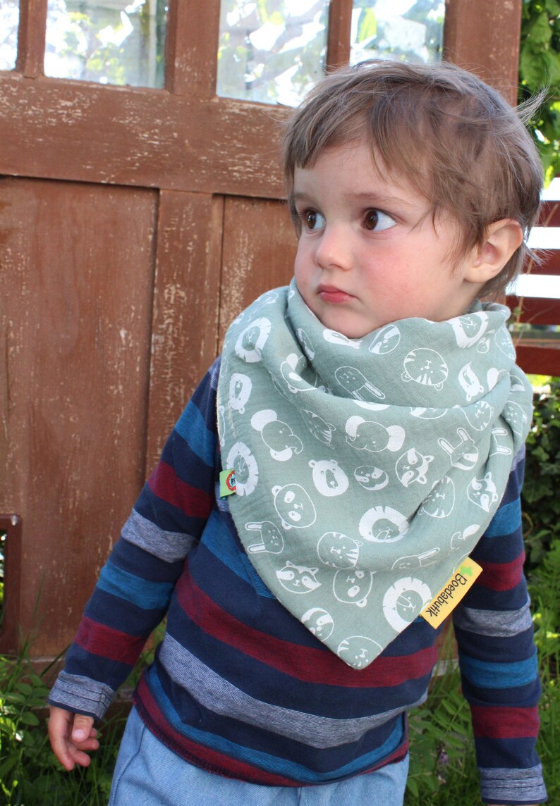 Puede incluir: Un ni&ntilde;o peque&ntilde;o con una camisa de manga larga a rayas azules y rojas y pantalones vaqueros azules. El ni&ntilde;o lleva una bandana verde con caras de animales blancas impresas. La bandana tiene una peque&ntilde;a etiqueta verde con la palabra "Boosdahik" impresa.