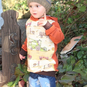 May include: A young child wearing a brown and blue striped knit hat, an orange and brown hooded sweatshirt with a tractor print, and blue jeans with a light blue pattern. The child is standing in front of a wooden post and green plants.