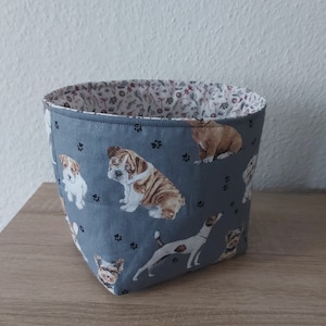 Könnte beinhalten: Ein grauer Stoffaufbewahrungskorb mit einer weißen und rosa floralen Auskleidung. Der Korb ist mit einem Muster verschiedener Hunderassen verziert, darunter eine Bulldogge, ein Terrier und ein Yorkshire Terrier.