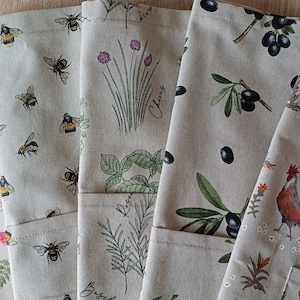Könnte beinhalten: Eine Sammlung beiger Küchenhandtücher aus Leinen mit verschiedenen Naturmotiven. Designs umfassen rote Mohnblumen, Bienen, Schnittlauch, Oliven und Hühner. Die Handtücher sind gefaltet und gestapelt, um die verschiedenen Muster zu zeigen.