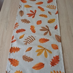 Könnte beinhalten: Rechteckiger Tischläufer mit einem Muster aus Herbstblättern in Orange-, Braun- und Gelbtönen auf hellgrauem Hintergrund. Die Blätter sind in verschiedenen Formen und Größen gehalten und schaffen ein saisonales Thema.