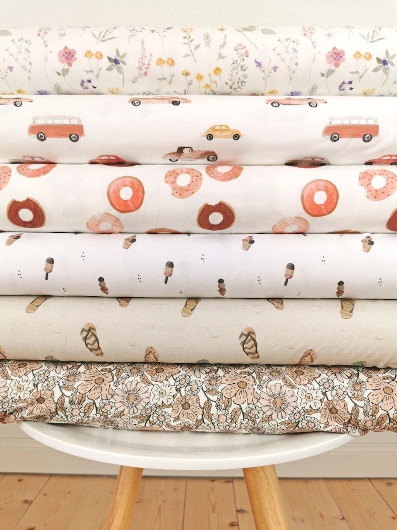 May include: A stack of six different fabric swatches with various patterns, including floral, cars, donuts, ice cream cones, sandals, and a floral pattern. The swatches are stacked on a white stool with wooden legs.