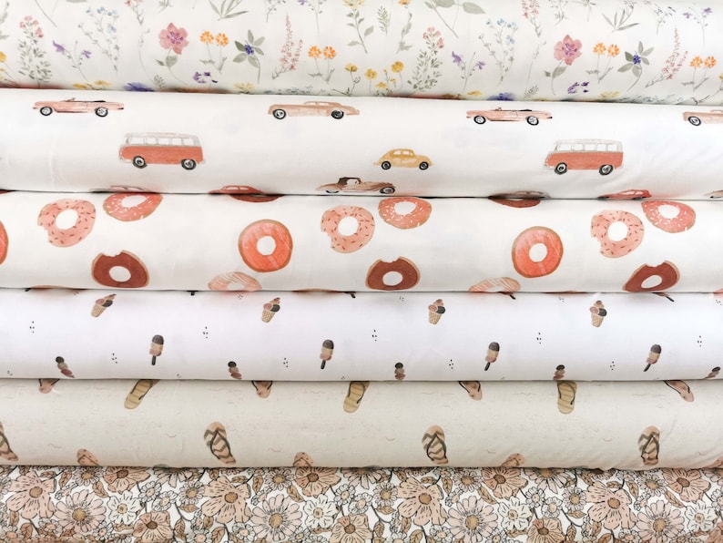 May include: Six rolls of fabric with different patterns. The top roll has a white background with small pink, yellow, and green flowers. The second roll has a white background with red and yellow cars and a red van. The third roll has a white background with pink donuts. The fourth roll has a white background with brown ice cream cones. The fifth roll has a white background with brown sandals. The bottom roll has a white background with small pink, yellow, and green flowers.