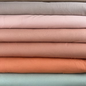 Könnte beinhalten: Ein Stapel verschiedenfarbiger Rippstrickstoffrollen. Die Farben umfassen hellbeige, beige, mauve, rosa, pfirsich, orange, meergrün und hellblau. Der Stoff hat eine vertikale Rippenstruktur.