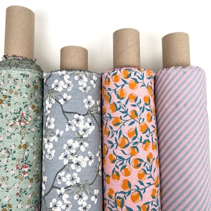 Könnte beinhalten: Vier Stoffrollen mit verschiedenen Mustern. Die erste Rolle hat einen grünen Hintergrund mit kleinen weißen und braunen Blumenmustern. Die zweite Rolle hat einen hellblauen Hintergrund mit weißen Blumenmustern. Die dritte Rolle hat einen rosa Hintergrund mit orangefarbenen Zitrusfrüchten. Die vierte Rolle hat ein rosa-graues Streifenmuster.