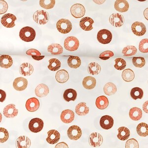 May include: White fabric with a repeating pattern of pink, brown, and gold donuts. The donuts have sprinkles and chocolate frosting.
