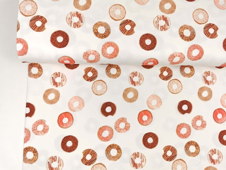 May include: White fabric with a repeating pattern of pink, brown, and gold donuts. The donuts have sprinkles and chocolate frosting.