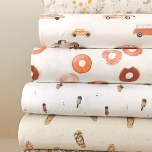 May include: A stack of six fabric rolls with different patterns. The top roll has a white background with small pink, yellow, and green flowers. The second roll has a white background with red, yellow, and blue cars. The third roll has a white background with orange donuts. The fourth roll has a white background with brown ice cream cones. The fifth roll has a beige background with brown flip flops. The bottom roll has a white background with pink and brown flowers.
