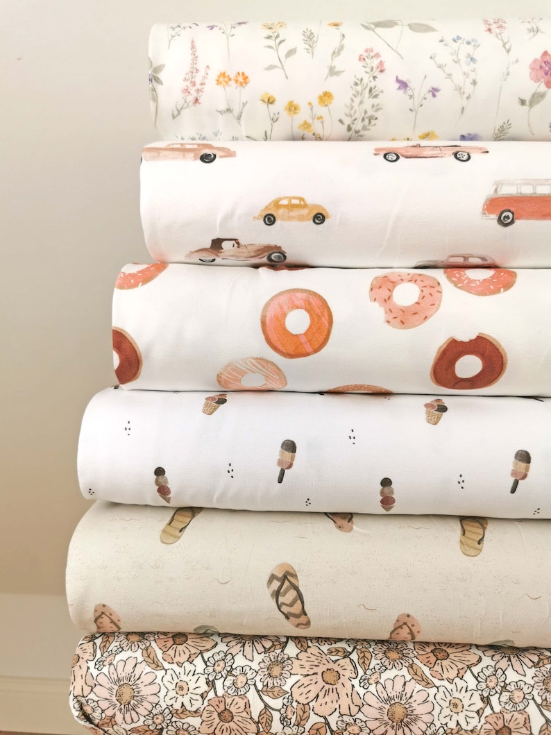 May include: A stack of six fabric rolls with different patterns. The top roll has a white background with small pink, yellow, and green flowers. The second roll has a white background with red, yellow, and blue cars. The third roll has a white background with orange donuts. The fourth roll has a white background with brown ice cream cones. The fifth roll has a beige background with brown flip flops. The bottom roll has a white background with pink and brown flowers.