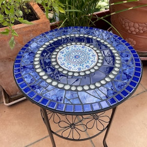 Könnte beinhalten: Ein runder Beistelltisch mit einer Mosaikplatte aus dunkelblauen Fliesen, einem zentralen runden Keramikmedaillon mit blau-weißem Blumenmuster und einem Rand aus kleinen weißen Glasperlen. Der Tisch hat ein schwarzes Metallgestell.