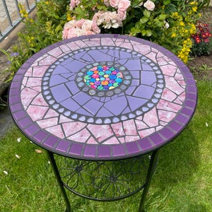 Peut inclure: Table d'appoint ronde en mosaïque avec un cadre en métal noir. Le plateau présente un motif en mosaïque avec des nuances de violet, de rose et un centre de pièces de verre colorées. La table mesure environ 51 cm de diamètre.