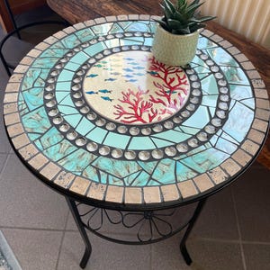 Könnte beinhalten: Runder Mosaik-Beistelltisch mit schwarzem Metallrahmen. Die Tischplatte hat ein türkisfarbenes und beigefarbenes Mosaikdesign mit einem zentralen Bild von Fischen und Korallen. Auf dem Tisch steht eine kleine grüne Topfpflanze.