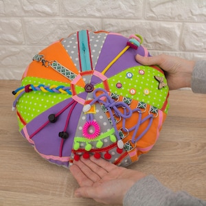 May include: A multi-textured sensory toy with a circular, segmented design. It features a variety of interactive elements, including a zip, buttons, and ribbons. The toy is made of fabrics in orange, green, purple, and grey. A small wooden butterfly is also visible.