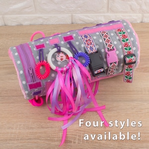 May include: A gray and white fabric cylinder with various textures and colors, including pink, purple, and red. It features a zipper, a buckle, ribbons, and a ring. The text "Four styles available!" is visible at the bottom of the image.