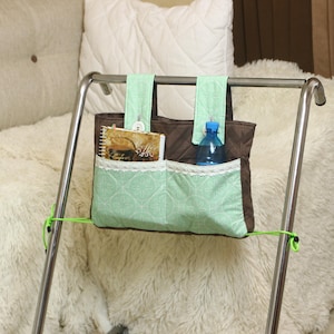 May include: A green and brown fabric organizer with pockets hangs on a metal bar. The organizer has a zippered top and two side pockets. A book and a water bottle are in the pockets.