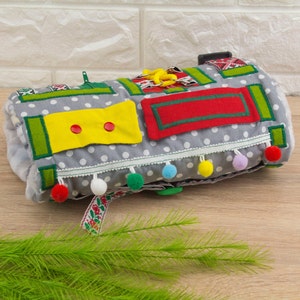 May include: A grey and white fabric activity toy with various textures and colours. It features a yellow button, red and green fabric squares, and a white lace trim. The toy is decorated with colourful pom-poms.