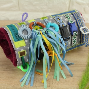 May include: A gray and white fabric activity mat with various textures and colors. It features a zipper, a buckle, a wooden ring, a plastic ring, and multiple ribbons in blue, yellow, and green.