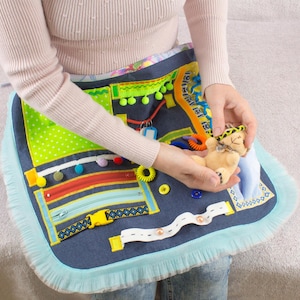 Puede incluir: Una alfombra de actividades sensoriales de mezclilla azul con varias texturas y colores. La alfombra presenta una cremallera, una hebilla, una cinta blanca, una cinta con estampado amarillo y negro, una tela de lunares verde y blanca, una tela a rayas roja y amarilla, una tela amarilla y verde con pompones, una tela blanca con un camino de pequeños botones blancos y un pequeño animal de peluche.