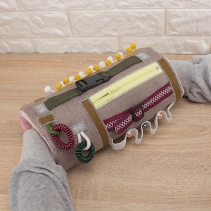 Puede incluir: Un juguete sensorial de tela marrón con varias texturas, incluyendo una cremallera, una hebilla y lazos de tela. El juguete está decorado con pompones amarillos y una cinta blanca.