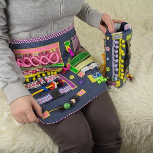 Op de afbeelding: Een spijkergoed schort met diverse kleurrijke bevestigingen, waaronder knopen, linten en kralen. Een bijpassende spijkergoed cilinder met knopen en pompons wordt in de buurt gehouden. De schort en cilinder zijn ontworpen voor sensorisch spel en leren.