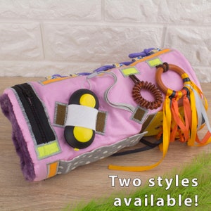 May include: A pink and purple fabric sensory toy with a zipper, a yellow button, a brown spiral, a wooden ring, and colorful ribbons. Two styles available.