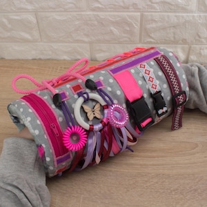 Puede incluir: Un juguete sensorial gris y rosa con varias texturas y actividades para niños. Cuenta con una cremallera, una hebilla, un anillo, una mariposa, cintas y una variedad de otros elementos sensoriales.