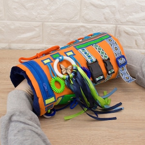 May include: Orange and blue fabric sensory toy with various textures, including ribbons, buckles, and a zip. The toy also features a wooden butterfly and a plastic ring.