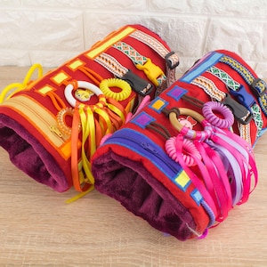 Puede incluir: Dos rollos de actividad sensorial de tela roja con varias texturas y colores. Un rollo tiene cintas amarillas, naranjas y blancas, una cremallera y una hebilla de plástico. El otro rollo tiene cintas rosas, moradas y azules, un anillo de madera y una hebilla de plástico azul.