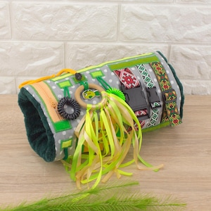 May include: A grey and green sensory activity mat with various textures and colours. It features a zipper, a wooden ring with a fish design, a rubber band, a plastic ring, and a set of yellow and green ribbons.