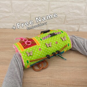 May include: A green and white polka-dotted sensory toy with wooden butterfly accents, a buckle, and wooden rings. The toy has the text "+Free Name Embroidery". The toy is designed for sensory exploration and development.