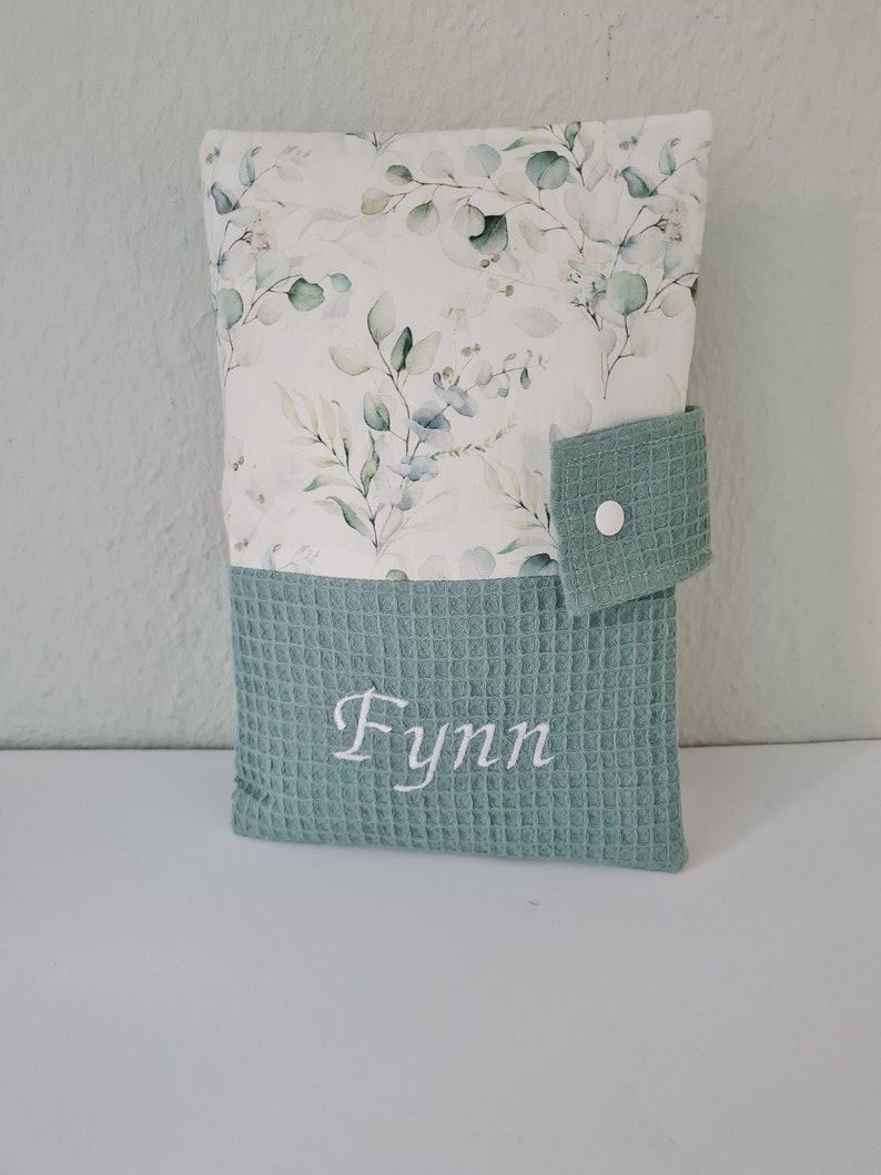 May include: A rectangular baby changing pad cover. The top portion features a white background with a green and blue floral pattern. The bottom is a textured, light green fabric with the name "Fynn" embroidered in white. A light green flap with a snap closure is on the right side.