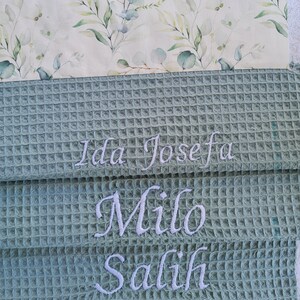 May include: A stack of three personalized waffle weave towels. The top towel has a floral pattern with green and blue leaves. The bottom three towels are a light green color with embroidered names: Ida Josefa, Milo, and Salih.