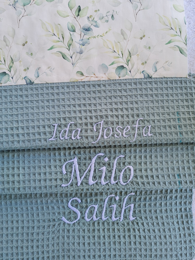 May include: A stack of three personalized waffle weave towels. The top towel has a floral pattern with green and blue leaves. The bottom three towels are a light green color with embroidered names: Ida Josefa, Milo, and Salih.