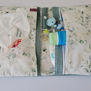 May include: A baby changing pad with a floral pattern of green leaves on a white background. The pad is open, revealing a pocket with tissues, a pacifier, a tube of lotion, and a diaper. The pad has a zipper closure and a teal waffle-weave trim.