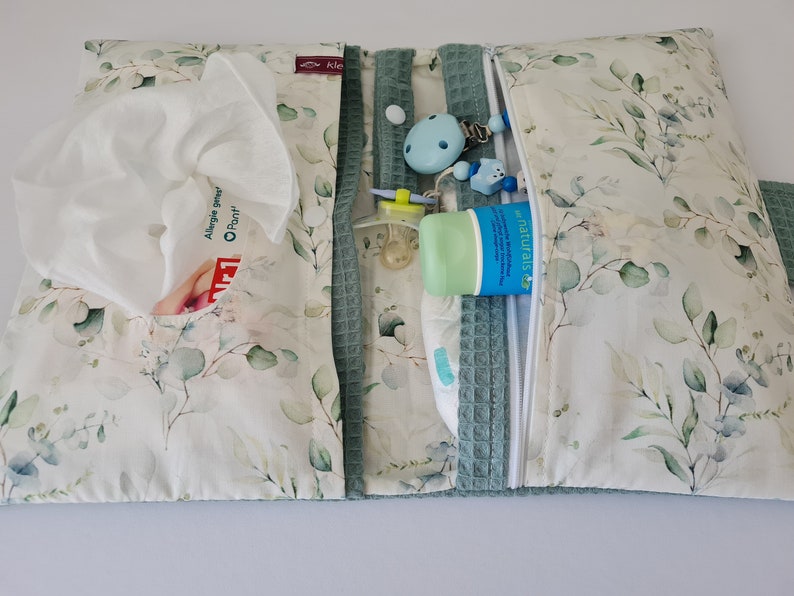 May include: A baby changing pad with a floral pattern of green leaves on a white background. The pad is open, revealing a pocket with tissues, a pacifier, a tube of lotion, and a diaper. The pad has a zipper closure and a teal waffle-weave trim.