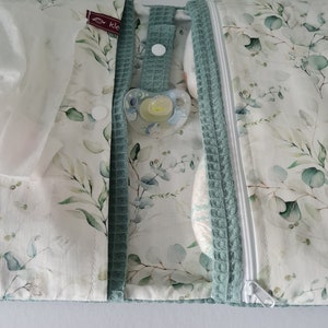 May include: A baby changing pad with a white and green eucalyptus leaf pattern. The pad has a teal waffle-textured trim and a white zipper. A pacifier is attached to the pad with a teal strap. The pad is open, revealing a diaper and wipes.