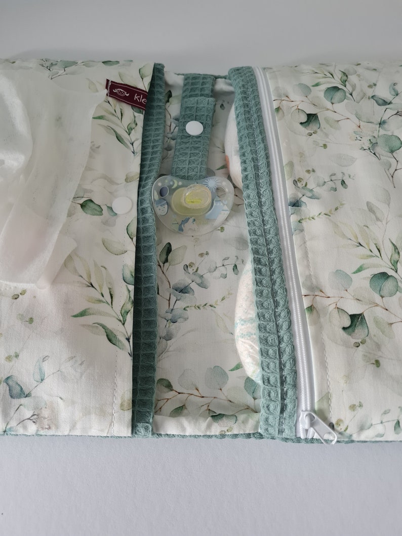 May include: A baby changing pad with a white and green eucalyptus leaf pattern. The pad has a teal waffle-textured trim and a white zipper. A pacifier is attached to the pad with a teal strap. The pad is open, revealing a diaper and wipes.