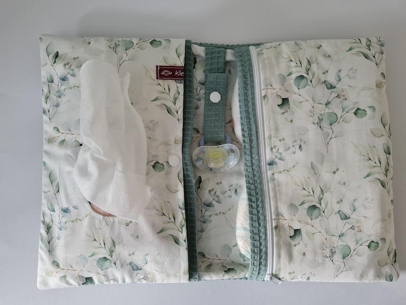 May include: A baby changing pad with a white and green eucalyptus leaf pattern. The pad is open, revealing a compartment for wipes, a pacifier, and a diaper. A green waffle-textured border and zipper are visible.
