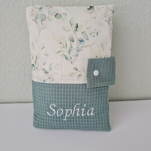May include: A baby changing pad cover with a floral pattern and the name "Sophia" embroidered in white. The top portion features a cream-colored fabric with green and blue leaves. The bottom is a textured, teal-colored fabric.