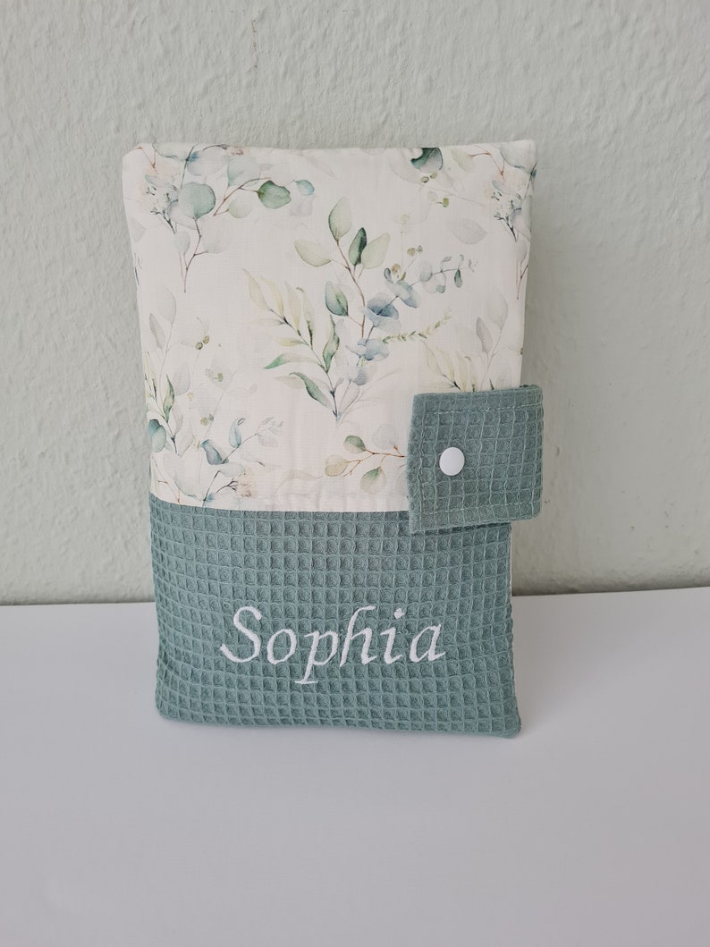 May include: A baby changing pad cover with a floral pattern and the name "Sophia" embroidered in white. The top portion features a cream-colored fabric with green and blue leaves. The bottom is a textured, teal-colored fabric.