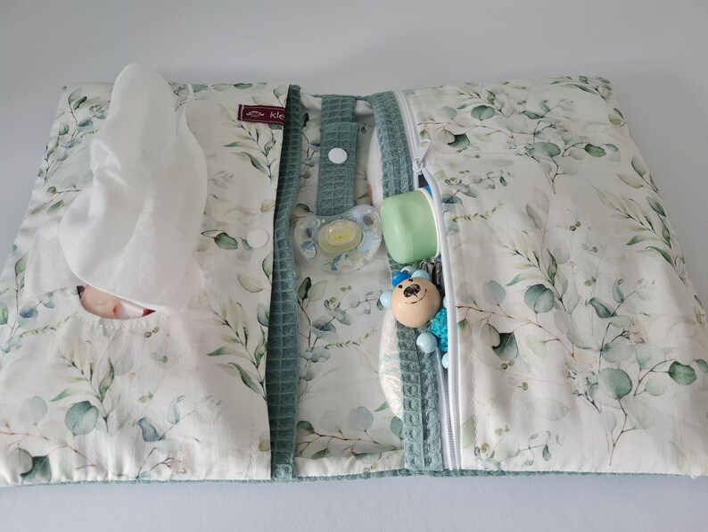 May include: A baby changing pad with a white and green eucalyptus leaf pattern. The pad has a pocket for wipes, a pacifier, a small toy, and a zippered compartment. The pad is made of soft fabric and has a teal waffle-textured trim.